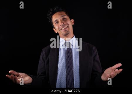 Sehr glücklicher Geschäftsmann mit offenen Armen, isoliert auf schwarzem Hintergrund. Der junge Geschäftsmann will sich umarmen. Junger lateinischer Mann freut sich über Bis zum nächsten Mal. Stockfoto