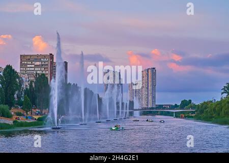 Die hohen Wasserstrahlen der Rusanivsky Fountains liegen am gleichnamigen Kanal, umgeben von einem üppigen grünen Park und Wohnhochhäusern, Kiew, Ukraine Stockfoto