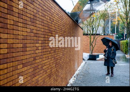 Amsterdam, Niederlande, 02/11/2021, Eine Frau, die einen Regenschirm hält, liest die Namen auf den Ziegelsteinen, an der Gedenkstätte.70 Jahre nach dem Zweiten Weltkrieg haben mehr als 102,000 Opfer des Holocaust endlich ihr eigenes Denkmal. Dieses nationale Denkmal befindet sich im Herzen des jüdischen Viertels von Amsterdam. Das Denkmal besteht aus einem Labyrinth von Durchgängen, flankiert von zwei Meter hohen Ziegelwänden, die die Botschaft ‘in Erinnerung an' vermitteln. Auf jedem der Ziegel ist der Name jedes Holocaust-Opfers eingeschrieben. Die meisten der Menschen, die das Denkmal besuchen, halten Papiere mit dem n Stockfoto