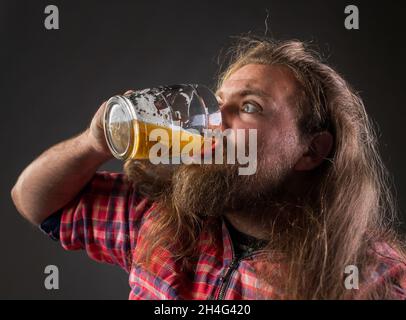 Unordentlicher Mann mit langen Haaren und Bart, der Bier aus dem Becher im Studio trinkt Stockfoto