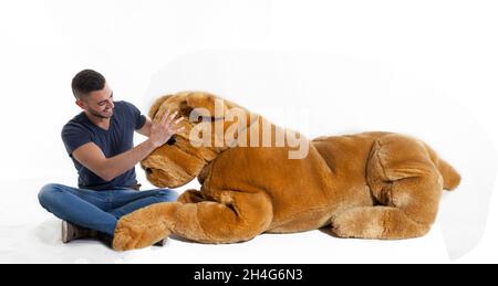 Junger Erwachsener, der mit einem riesigen Spielzeug-Hund auf weißem Hintergrund sitzt - Studioaufnahme Stockfoto