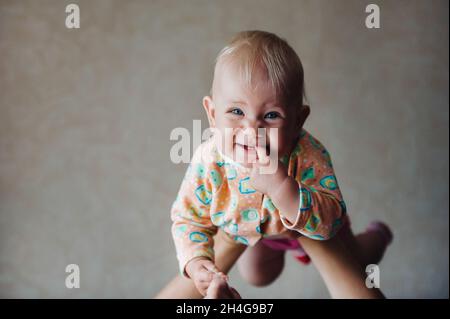 Ein kleines Mädchen in den Armen ihrer Mutter über ihrem Kopf lächelt mit einem Finger im Mund. Stockfoto