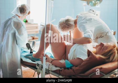 Im Krankenhaus gibt die Hebamme einer Mutter ein Neugeborenes Baby, den unterstützenden Vater, der in der Nähe der Frau sitzt, zum Halten. Glückliche Familie in der modernen Delivery ward Stockfoto