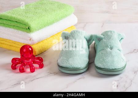 Rotes Spa-Massagegerät und lustige, gemütliche Hausschuhe in der Nähe von bunten Frottee-Handtüchern auf der Marmoroberfläche. Massagegerät, warme Hausschuhe und Baumwollpflegeartikel. Stockfoto