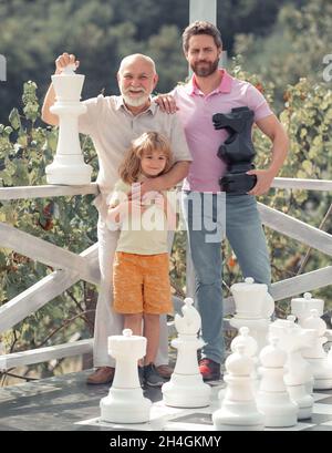 Großvater mit Sohn und Enkel spielen Schach. Erstaunte Männer in verschiedenen Altersgruppen spielen Schachbrett Spiel. Drei-Mann-Generation. Väter Tag Konzept. Stockfoto