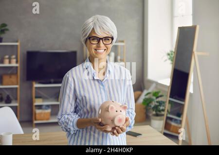 Porträt einer glücklichen älteren Frau, die mit ihren Ersparnissen und ihrem Lächeln ein Sparschwein in der Hand hält Stockfoto