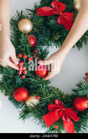 Draufsicht auf Kinderhände, die den Weihnachtskranz mit roten Weihnachtsspielzeugen und Schleifen schmücken. Advent für Kinder, DIY. Stockfoto