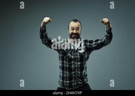 Halbkörperporträt eines bärtigen Mannes im Karomudio, der im Triumph die Hände hebt, Studiohintergrund. Stockfoto