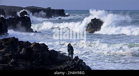Zwei Menschen, die an der schwarzen Lavaküste mit Blick auf die stürmische Brandung standen, umarmten Skardsvik, Snaefellsnes, Island Stockfoto