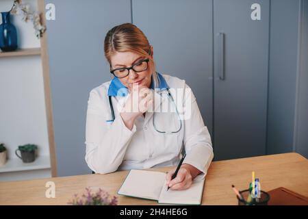 Krankenschwester mittleren Alters in weißen Kleidern sitzt an einem Schreibtisch im Krankenhaus und schreibt etwas in ein Notizbuch. Vorderansicht. Stockfoto