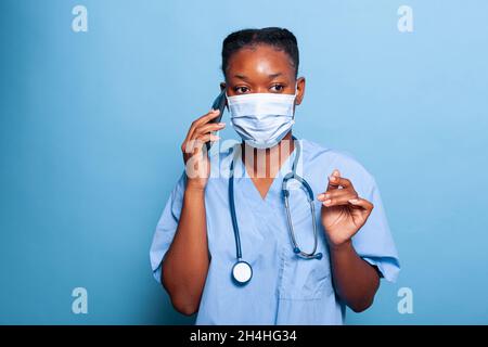 afroamerikanischer Praktikerassistent mit Schutzmaske gegen Coroanvirus, der mit einem Kollegen am Telefon telefoniert und die Erfahrung mit Krankheiten während der Arbeit im Studio erklärt. Medizinkonzept Stockfoto