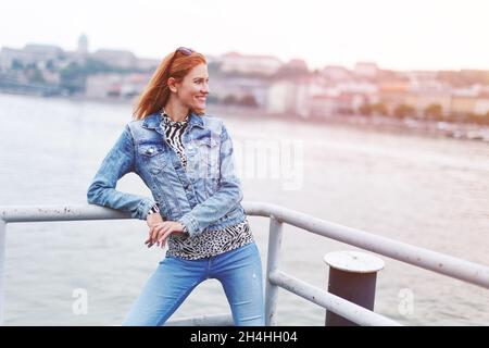 Junge Rotschopf kaukasischen modische Frau toothy lächeln am Flussufer in Sonnenuntergang Stockfoto