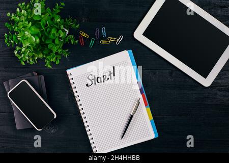 Ansicht des offenen Notizbuchs mit STARTBESCHRIFTUNG. Grüne Blume, Tablet, farbige Büroklammern, smart, Stift und Notizbuch auf Holzhintergrund. Stockfoto