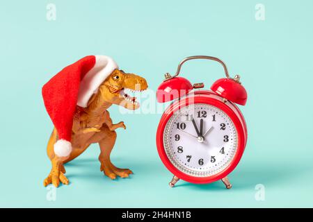 Dinosaurier Rex in rotem Weihnachtsmann Hut und Wecker auf blauem Hintergrund Silvester oder Heiligabend Art Weihnachtskarte Kreative Idee für Merry Xmas Co Stockfoto