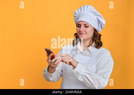 Happy Woman Chef Blick auf iphone auf gelben Studio-Hintergrund, Copy Space - Moskau, Russland, 20. Oktober 2021 Stockfoto