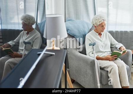 Hochwinkelporträt einer weißhaarigen älteren Frau, die zu Hause Buch liest, während sie im Sessel sitzt, mit Spiegelung aufgenommen, Platz zum Kopieren Stockfoto