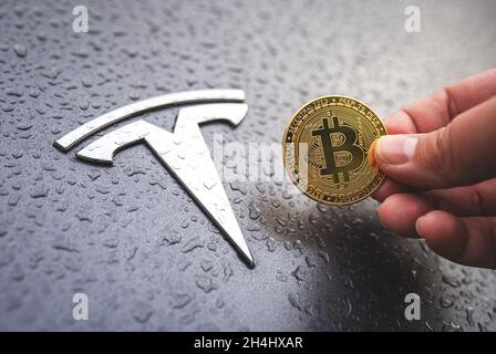 Bitcoin-Münze in der Hand mit Tesla-Auto-Logo auf dem Hintergrund. Nahaufnahme, selektiver Fokus. Tesla akzeptiert das Kryptowährungskonzept von Bitcoin. Warschau, Polen - 23. Oktober 2021 Stockfoto