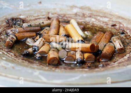 Viele Zigarettenstummel in einem grauen Standaschenbecher, draußen stehend, in den es hinein gegnet hat. Stockfoto