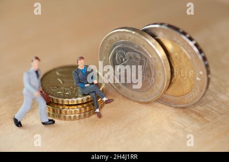 Makro aus kleinen Figuren, die auf einem Münzstapel daneben sitzen, sind zwei zwei Euro-Münzen mit einem Symbol für Griechenland Stockfoto