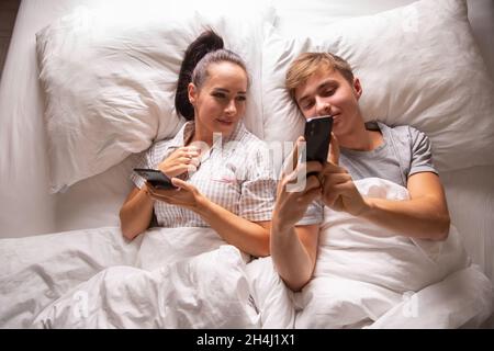 Draufsicht auf ein Paar, das interessante und lustige Dinge auf ihren Mobiltelefonen teilte, während es vor dem Schlafen in weißer Bettwäsche lag. Stockfoto