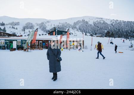 Kalavryta Ski Resort, Griechenland - 1. Januar 2020: Ältere schöne Frau im langen grauen Mantel im Skiresort-Verleih mit schneebedeckter Aussicht auf die Berge im Hintergrund. Winterurlaub Stockfoto