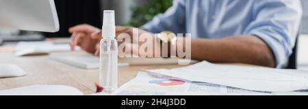Beschnittene Ansicht des Händedesinfektionsmittels in der Nähe eines verschwommenen afroamerikanischen Geschäftsmannes, der Computer, Banner verwendet Stockfoto