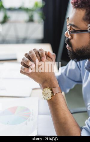 Seitenansicht des afroamerikanischen Geschäftsmannes in Brillen, der in der Nähe verschwommener Papiere im Büro sitzt Stockfoto