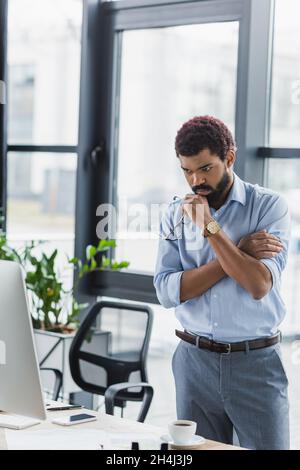 Fokussierter afroamerikanischer Geschäftsmann, der im Büro eine Brille in der Nähe des Computermonitors hält Stockfoto