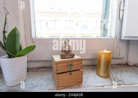 Goldene Zen-Buddha-Statue, die neben dem Fenster mit Pflanzen meditiert. Kerzenlicht und Truhe im Hintergrund. Stockfoto