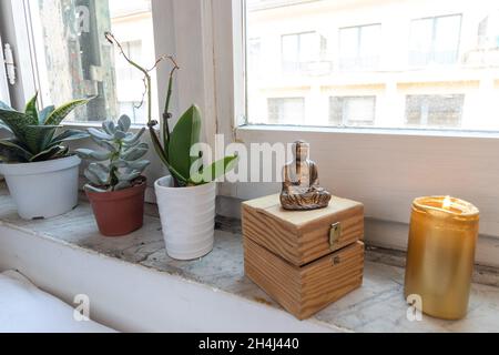 Goldene Zen-Buddha-Statue, die neben dem Fenster mit Pflanzen meditiert. Kerzenlicht und Truhe im Hintergrund. Stockfoto