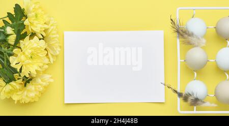 Ostereier hellblau in weißem Vintage Metallhalter mit Federn, Band, gelben Chrysanthemen Blumen und leeres Papier für Text auf gelbem Hintergrund Stockfoto