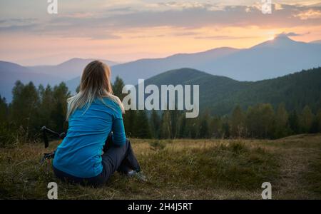 Rückansicht einer blonden jungen Frau, die auf einem grasbewachsenen Hügel sitzt und einen malerischen Blick auf den Sonnenaufgang in den Bergen genießt. Weibliche Wandererin, die sich auf Gras in der Nähe von Trekkingstöcken ausruht. Konzept von Reisen, Wandern, Natur. Stockfoto