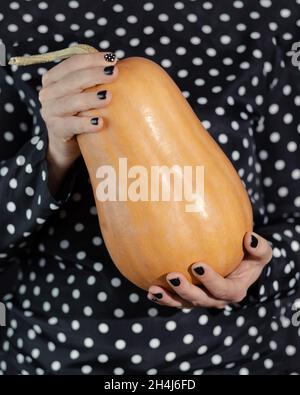 Eine Frau in einem schwarzen Kleid mit weißen Tupfen hält einen schönen orangefarbenen Kürbis in den Händen. Minimalismus, modisches Halloween. Stockfoto