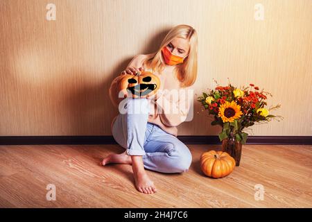 Unglückliche junge blonde Frau, die eine medizinische Maske in einem warmen Pullover trägt, sitzt auf dem Boden und hält einen Kürbis in den Händen, einen herbstlichen Strauß mit einer Sonnenbräunung Stockfoto
