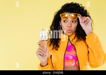 Überrascht afroamerikanische junge Frau in Sonnenbrillen halten Eis-Kegel isoliert auf gelb Stockfoto