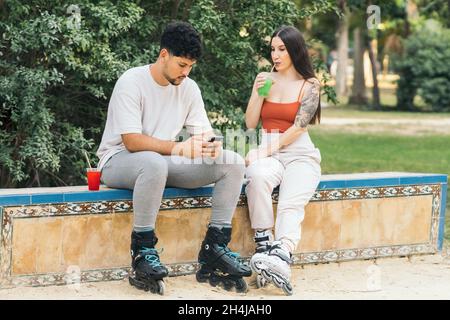 Ein Paar nutzt ein Handy, während es in einem Park mit Inline-Skates ein alkoholfreies Getränk trinkt Stockfoto