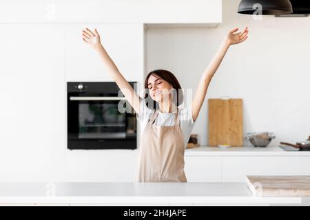 Entspannte junge Dame, die die Hände hebt und sich begeistert fühlt, allein in der modernen Kücheneinrichtung zu kochen und Spaß zu haben Stockfoto