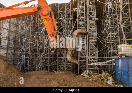 Orangefarbener Greifer mit Baggerschaufel vor einem Hochregallager, das abgerissen wird Stockfoto