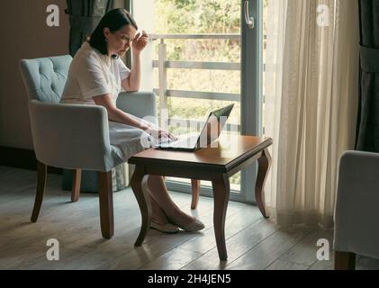 Frau, die im Heimbüro einen Laptop benutzt. Eine attraktive Frau mittleren Alters sitzt mit einem Laptop in der Nähe des Fensters und arbeitet mit Blick auf den Bildschirm Stockfoto