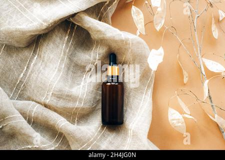Braune Glas-Serumflasche auf Textil-beigen Hintergrund mit Lunaria trockenen Blüten. Konzept der Naturkosmetik. Draufsicht, flach liegend. Stockfoto