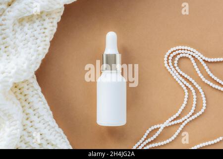 Weiße Serumflasche auf beigem Hintergrund mit Strickstoff und Perlenkette. Konzept der Naturkosmetik. Draufsicht, flach liegend. Stockfoto