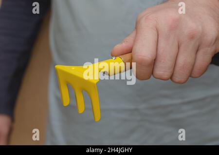 Gartengeräte für Kinder. Die Hand eines Mannes hält einen Spielzeugrechen. Gelber Rechen mit Holzgriff. In Innenräumen. Selektiver Fokus. Stockfoto