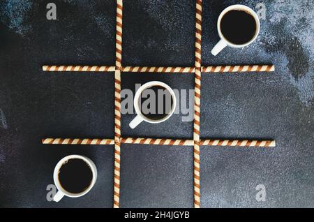 Geschäftsmann arbeitet morgens mit Tassen heißen Kaffee und süßen Croissants auf schwarzem Hintergrund. Spiel von Nullen und Kreuzen. TIC tac TOE. Stockfoto