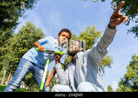 Schöne glückliche afroamerikanische Familie im Park - Schwarze Familie, die Spaß im Freien hat, Vater, der Sohn lehrt, auf dem Roller zu fahren Stockfoto