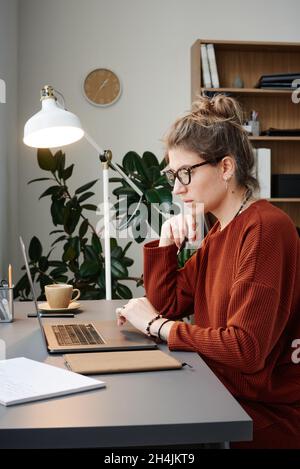 Seriöse junge Frau, die sich auf ihre Online-Arbeit konzentriert, während sie am Tisch sitzt und am Laptop arbeitet Stockfoto
