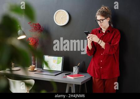 Junge Frau in legerer Kleidung liest eine Nachricht auf ihrem Mobiltelefon, während sie neben dem Tisch mit einem Laptop gegen die schwarze Wand steht Stockfoto