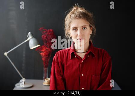 Porträt einer hübschen jungen Frau im roten Hemd, die auf die Kamera schaut, die in einem modernen Zimmer mit Arbeitsplatz im Hintergrund steht Stockfoto