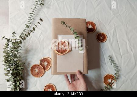 Ansicht von oben einer Person, die ein Paket in braunem Papier mit einem leeren Umschlag und rustikalen Eukalyptus- und Orangendekorationen hält Stockfoto