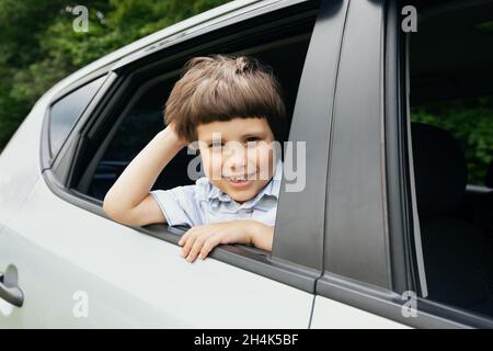 Nahaufnahme eines fröhlichen kleinen Jungen, der während einer Fahrt aus dem Autofenster schaut, Porträt eines entzückenden kleinen männlichen Kindes, das Autofahrten genießt, Stockfoto