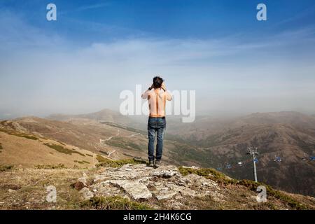 Ein junger Mann, von hinten gesehen, steht auf dem Gipfel eines Berges, um mit einem Fernglas die Aussicht zu betrachten. Stockfoto